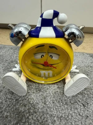 Gul M&M väckarklocka med mössa - Rolig väckarklocka formad som en gul M&M-figur med blåvit randig sovmössa och silvriga klockklockor. Klockan har vita armar och ben samt stora vita skor. Perfekt som dekoration eller för att vakna med stil.