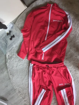 Rött tracksuit set Palm Angels - Säljer ett rött tracksuit set från Palm Angels med vita ränder längs ärmar och ben. Setet består av en zip-jacka och matchande byxor, båda med Palm Angels-logga broderad framtill. Perfekt för en sportig känsla. Perfekt för en streetwear-look.