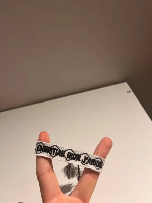 Svartvitt armband Christian Dior - Snyggt svartvitt tygarmband från Christian Dior med broderad logga och dekorativa tofsar i ändarna. Armbandet har ett grafiskt mönster och knyts enkelt runt handleden. Perfekt accessoar för att lyfta din outfit.