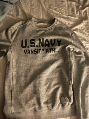 Grå sweatshirt U.S. Navy Champion - Grå sweatshirt från Champion med tryck 'U.S. NAVY VARSITY ATHL.' på bröstet. Klassisk crewneck-modell med ribbade muddar och en patch nertill. Perfekt för en avslappnad och sportig stil.