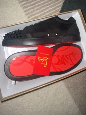 Svarta Louboutin sneakers med nitar - Svarta sneakers i mocka från Christian Louboutin med ikoniska röda sulor och svarta nitar på tån. Låga i modellen och detaljerade med metallnitar på sidan. Perfekta för dig som vill sticka ut med en lyxig och edgy stil.
