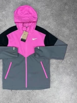 Nike vindjacka i rosa och grått - Snygg vindjacka från Nike i färgerna rosa, grå och svart. Jackan har huva, dragkedja framtill och två fickor med rosa dragkedjor. Perfekt för blåsiga dagar och sportiga outfits. Nike-logga på bröstet och modern passform.