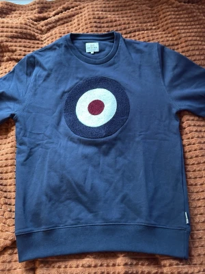 Mörkblå sweatshirt med rundel - Säljer en mörkblå sweatshirt med ett stort rundel-motiv i vitt, marinblått och rött på bröstet. Tröjan har rund halsringning och långa ärmar. Tillverkad i mjuk bomull för skön känsla och avslappnad stil.