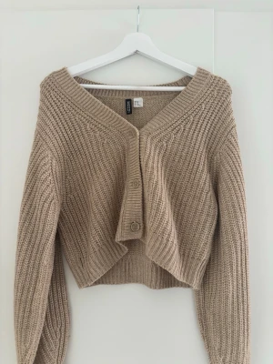 Beige stickad kofta från H&M Divided - Säljer en beige stickad kofta från H&M Divided i storlek XS. Koftan har en croppad passform, grovstickad struktur och två stora knappar framtill. Perfekt till lager på lager och har bred ribbad kant vid ärmslut och nederkant.