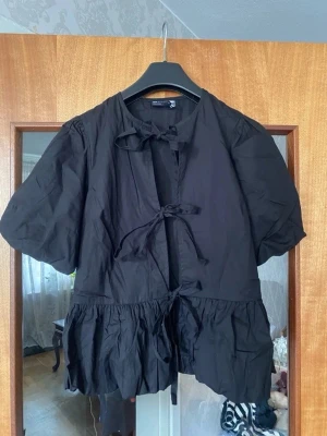Svart volangblus med knytband - Trendig svart blus med puffiga korta ärmar och volangkant nedtill. Blusen har två knytband framtill och en avslappnad passform. Perfekt för dig som gillar en lite mer edgy och unik stil. Tillverkad i ett lätt tyg som ger en luftig känsla.