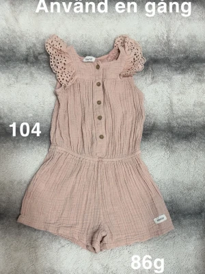 Rosa jumpsuit med spetsärmar - Supersöt rosa jumpsuit i mjukt, luftigt tyg med knappar framtill och korta ben. Ärmarna har fina spetsdetaljer som ger en gullig touch. Perfekt för varma dagar och enkel att ta på. Liten etikett nertill på benet.🌸Använd en gång. Storlek 104