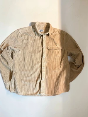 Beige manchesterskjorta från Weekday - Säljer en beige manchesterskjorta från Weekday med smala vertikala ränder. Skjortan har klassisk krage, bröstficka och knäppning framtill. Perfekt för dig som gillar en avslappnad men ändå stilren look.