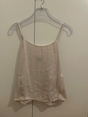 Beige linnetopp med volang - Säljer en ljusbeige linnetopp med tunna axelband och volangdetalj baktill. Toppen har en lös passform och är tillverkad i ett svalt material som passar perfekt till sommaren. Enkel och stilren design som är lätt att matcha. Lite sönder vid ena toppens band 