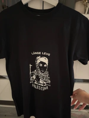 Svart t-shirt med Palestina-tryck - Svart t-shirt med vitt tryck framtill som föreställer en person i sjal och texten 'Länge leve Palestina'. Klassisk rund hals och korta ärmar. Perfekt för dig som vill göra ett statement med din stil.