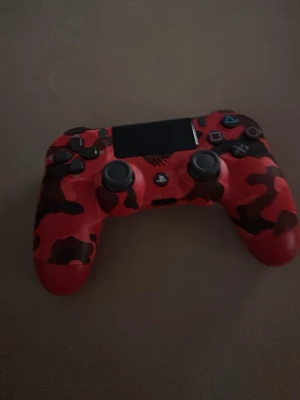Röd kamouflage PS4 handkontroll - Säljer en PlayStation 4 handkontroll från Sony med röd och svart kamouflagemönster på framsidan och helröd baksida. Kontrollern har klassisk DualShock 4-design med touchpad, analogspakar och knappar i plast. Perfekt för gaming och snygg i handen.