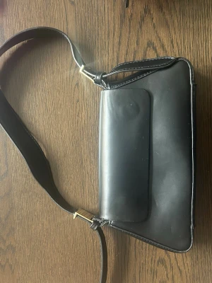 Svart axelväska Zara - Stilren svart axelväska i slätt läder med justerbar axelrem och guldfärgade metalldetaljer. Väskan har en rektangulär form och ett lock framtill. Perfekt för dig som gillar minimalistisk och klassisk design.