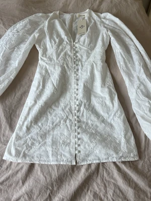 Vit Adoore klänning! - Superfin vit kortklänning från Adoore med broderade blommor, v-ringning och långa puffärmar. Klänningen har klädda knappar framtill och markerad midja för en snygg siluett. Perfekt för sommarens alla tillfällen! Och midsommar!🌸helt ny, aldrig använd.