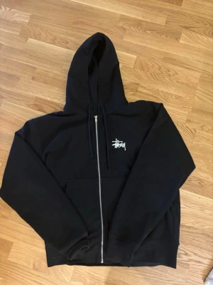 Svart zip hoodie från Stüssy - Säljer en svart hoodie från Stüssy med dragkedja framtill och stor vit logga på ryggen samt mindre logga på bröstet. Hoodien har huva med dragsko och en stor magficka. Perfekt för dig som gillar streetwear och vill ha en klassisk look.