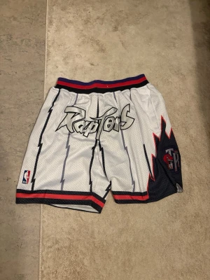 Toronto Raptors basketshorts - Säljer ett par vita basketshorts med Raptors-logga och coolt blixtmönster. Shortsen har röda och svarta detaljer, elastisk midja och meshmaterial för extra komfort. Perfekta för dig som gillar streetwear och NBA-stil. I perfekt skick bara att snöret runt midjan saknas, då jag själv har tagit ut den. Använd fåtal gånger. 