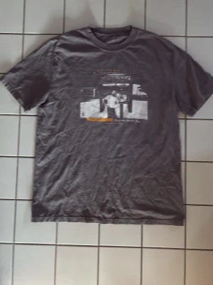 Vintage-look John Galt T-shirt – McCallaghan NYC Graphic 🎸 - Säljer nu denna snygga gråa t-shirt från John Galt (Brandy Melville). Den har ett coolt vitt  fototryck framtill med texten "McCallaghan – The Golden Hearted Boy / New York City"Stil: Washed/vintage look som passar perfekt till baggy jeans, shorts eller en kjol. Passar dig som gillar graphictee, streetwear, grunge och vintagestil. Mycket bra skick, inga synliga defekter. Strl L. Passform: Skön passform som passar både kille och tjej (unisex/oversize beroende på önskad stil)Material:100% bomull