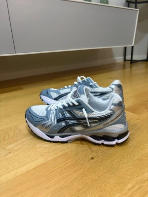 ASICS vita och grå sneakers - Snygga sneakers från ASICS i vitt och grått med svarta detaljer. Skorna har en sportig design med meshpaneler för bra ventilation och reflekterande inslag. Sulan är vit med svarta och grå partier för extra grepp. Perfekta för en trendig och aktiv look.