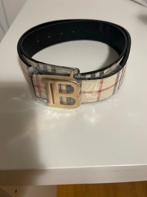Rutig Burberry bälte med B-spänne - Snyggt bälte från Burberry med klassiskt rutigt mönster i beige, svart och rött. Bältet har ett stort guldfärgat B-spänne och insidan är svart. Perfekt accessoar för att lyfta din outfit. storleks PIN ingår