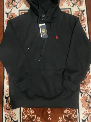 Svart hoodie från Polo Ralph Lauren - Svart hoodie från Polo Ralph Lauren med klassisk röd logga broderad på bröstet. Hoodien har huva med dragsko, magficka och ribbade muddar vid ärmar och nederkant. Tillverkad i mjukt bomullsmaterial som känns skönt mot huden. Det står M med det är passar perfekt till S 