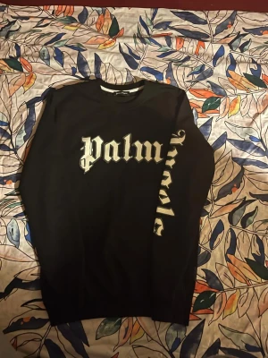 Svart Palm Angels sweatshirt - Svart sweatshirt från Palm Angels med stor vit logotyptext i gotisk stil över bröstet och ärmen. Tröjan har rund halsringning och ribbade muddar. Perfekt för dig som gillar streetwear och vill ha en statement piece i garderoben.