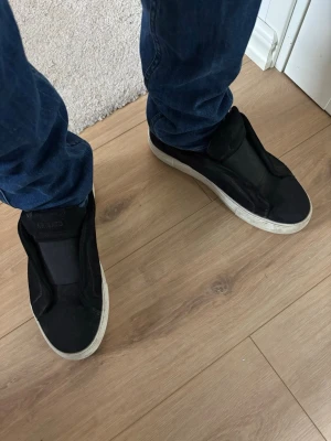 Svarta Arigato slip-on sneakers - Säljs inte längre. Svarta slip-on sneakers med vit sula och minimalistisk design. Ovandelen är i tyg och skon har en låg profil utan synliga snören. Perfekta för en avslappnad och stilren look.