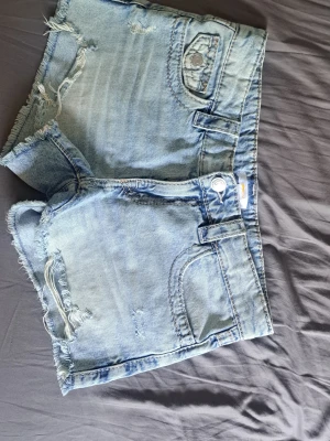 Gina Tricot ljusa shorts - Säljer ett par ljusblå jeansshorts med råa kanter och slitningar från Gina tricot. Shortsen har snygga bakfickor med broderade detaljer och knappstängning. Mått finns på bilden.