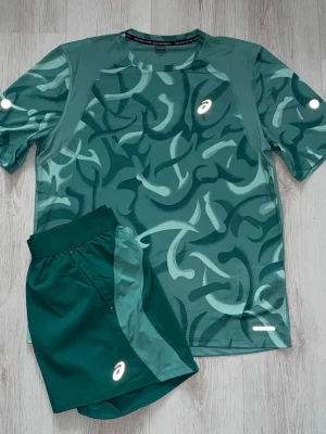 Grön träningsset från Asics - Snyggt träningsset från Asics med grön mönstrad t-shirt och matchande shorts. T-shirten har ett abstrakt mönster i olika gröna nyanser och reflekterande detaljer. Shortsen är mörkgröna med ljusare paneler och Asics-logga. Perfekt för löpning eller gym.