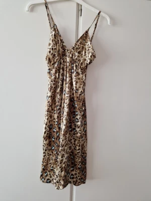 Leopardmönstrad satin klänning - Snygg leopardmönstrad klänning i satin med tunna axelband och v-ringad hals. Klänningen har en lätt glansig finish och ett avslappnat fall. Perfekt för dig som vill sticka ut med ett djurmönster och en feminin siluett.
