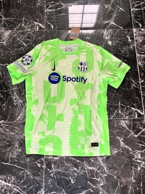 Barcelona Nike matchtröja neon/grön - Säljer en Barcelona matchtröja från Nike i neon/grön färg med digitalt mönster. Tröjan har klubbmärke, Nike-logga och Spotify som sponsor framtill samt Champions League-märke på ärmen. Tillverkad i lätt och ventilerande material, perfekt för fotboll.