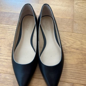 Svarta ballerinaskor från Lauren Ralph Lauren helt nya och oanvända  - Stilrena klassiska svarta ballerinaskor i skinn från Lauren Ralph Lauren. Skorna har spetsig tå och guldfärgad RL-logga bak på hälen. Klassisk design med låg klack och beige innersula för extra komfort. Stl 36. Helt nya och oanvända, säljes pga fel storlek. Nypris 1695kr inköpta april -26 på Ellos. Säljes för 1100kr