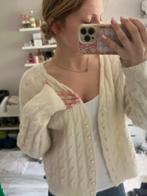 Krämvit kabelstickad kofta Vero Moda - Säljer en krämvit kabelstickad kofta från Vero Moda. Koftan har v-ringning, pärlknappar framtill och långa ärmar med ribbade muddar. Perfekt att slänga över ett linne för en chill och snygg look.