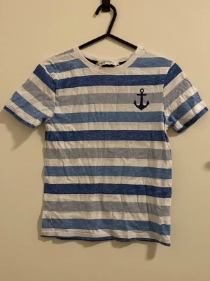 Randig t-shirt med ankare från H&M - Säljer en fräsch t-shirt från H&M med blå och vita ränder och ett tryckt ankare på bröstet. Perfekt för en chill och stilren look. T-shirten är kortärmad och gjord i mjuk bomull. Mått: från ena axel till den andra är ca 32cm och längden är 47cm