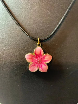 Halsband med rosa blomma - Unikt halsband med ett hänge i form av en fembladig blomma i rosa och vitt med gulddetaljer. Hänget sitter på ett svart rem som ger en snygg kontrast till den färgstarka blomman. Perfekt för dig som vill ha en söt och färgglad accessoar.