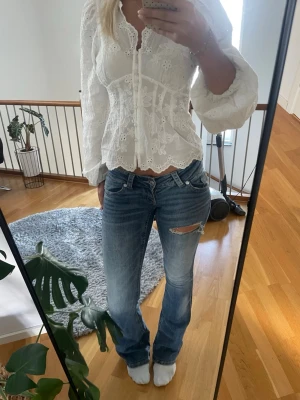 Blå slitna bootcut jeans - Säljer ett par blå bootcut jeans med slitningar och hål på låret. Jeansen har klassisk femficksdesign, låg midja och är i ett mjukt denimtyg. Perfekta för en avslappnad och trendig look.