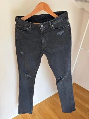 Svarta slitna jeans med raka ben - Säljer ett par svarta jeans med raka ben och diskreta slitningar vid knä och ficka. Jeansen har klassisk femficksmodell och är tillverkade i ett mjukt denimtyg. Perfekta för en avslappnad och trendig look.