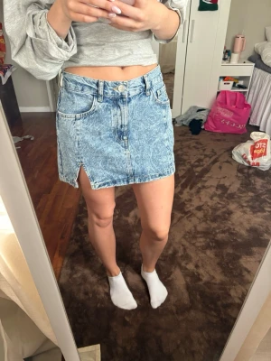 Blå jeanskjol med slits -  Säljer en snygg blå jeanskjol i kort modell med en liten slits framtill. Kjolen har klassisk femficksdesign och stängs med knapp och dragkedja. Perfekt till sommaren och enkel att matcha med olika toppar.