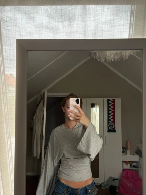 Grå croppad topp med vida ärmar - Säljer en ljusgrå croppad topp med supervida, långa ärmar. Toppen har en enkel och clean design utan synliga mönster eller detaljer, perfekt för en chill och trendig look. Materialet känns mjukt och stretchigt, troligen bomull eller polyester.