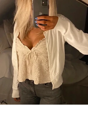 Krämvit linnetopp med spetsdetaljer - Söt krämvit linnetopp med smockad struktur och spetskant vid urringningen. Toppen har tunna axelband och en liten rosett framtill. Perfekt att styla med jeans eller kjol för en trendig look.