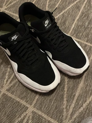 Nike Air Max svarta och vita sneakers - Nike Air Max sneakers i svart med vita detaljer och sula. Klassisk låg modell med perforerad tå, svarta skosnören och tydlig Nike-logga på sidan och plösen. Snygg och sportig design som passar till det mesta.