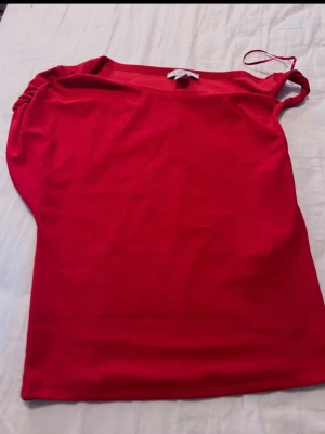 Röd oneshoulder topp med rynk - Snygg röd oneshoulder topp med rynkdetalj på axeln. Toppen är ärmlös och har en tight passform som framhäver figuren. Perfekt för dig som vill sticka ut med en enkel men trendig look.