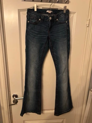 Blå bootcut jeans med slitningar - Säljer ett par blå bootcut jeans med snygga slitningar och lätt tvättade detaljer. Jeansen har fem fickor, knappgylf och utsvängda ben. Perfekta för dig som gillar en retro och avslappnad stil.