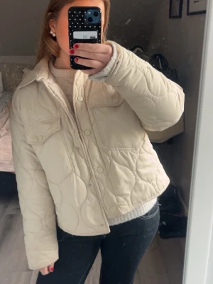 Beige quiltad pufferjacka - Säljer en beige pufferjacka med quiltat mönster och knappar framtill. Jackan har två bröstfickor med lock och en klassisk krage. Perfekt för kalla dagar och har en trendig, boxig passform. Materialet är mjukt och isolerande. Kan gå ner i pris vid snabbt köp! Lite skitig men lär gå bort i tvätten 
