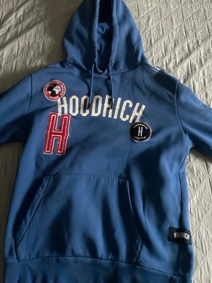 HOODRICH Hoodie Blå  - Snygg blå hoodie från Hoodrich med stor huva och dragsko. Framsidan har vita tryckta bokstäver, två patchar och en stor röd H-detalj. Känguruficka framtill och ribbade muddar. Materialet känns mjukt och bekvämt, perfekt för en avslappnad streetwear-look.