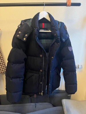 Mörkblå Ralph Lauren jacka  - Säljer en mörkblå pufferjacka med avtagbar huva och stora fickor framtill. Jackan har quiltad design, hög krage och knäppning med både dragkedja och knappar. Perfekt för kalla vinterdagar. Köpt för över 5000kr på Zalando kvitto och äkthets bevis finns för den som är intresserad. Jackan användes bara ett par gånger i vintras så inga täcken på användningen.