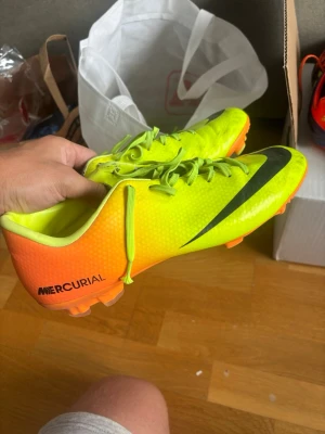 Nike Mercurial neon fotbollsskor - Säljer ett par Nike Mercurial fotbollsskor i neonfärg med svart swoosh och stor logga på hälen. Skorna har snörning, låg profil och är tillverkade i syntetmaterial för bra grepp och lätt känsla på planen. Perfekta för dig som vill sticka ut på fotbollsplanen.
