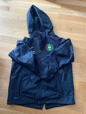 Svart Nike vindjacka FC Järfälla - Snygg svart vindjacka från Nike med FC Järfälla-logga på bröstet och initialerna JF som står för Järfälla fotboll. Jackan har huva, hel dragkedja och två sidofickor. Tillverkad i lätt och tåligt material, perfekt för blåsiga dagar.