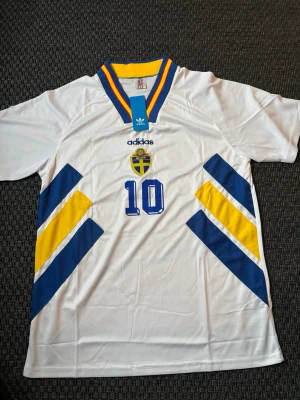 Svensk landslagströja Adidas nr 10 DAHLIN - Vit svensk landslagströja från Adidas med nummer 10 på bröstet, SVFF-emblem och blå/gula detaljer. Tröjan har V-ringad krage med blå och gula ränder. Perfekt för fotbollsfans som vill stötta Sverige.