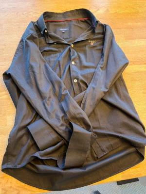 Svart skjorta från GANT i bomull - Klassisk svart skjorta från GANT i storlek L (41/42). Skjortan har vita knappar, broderad logga på bröstet och en snygg röd detalj på insidan av kragen. Tillverkad i mjuk bomull och har lång ärm med manschett.