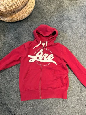 Röd hoodie med dragkedja från Årebutiken - Snygg röd hoodie från Årebutiken med vit broderad text 'Åre' och detaljer om Jämtland, Sweden. Tröjan har dragkedja framtill, två fickor och justerbar huva med vita snören. Perfekt för dig som gillar en sportig och avslappnad stil. 