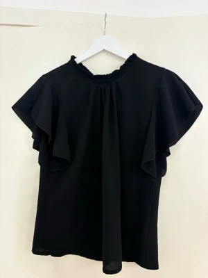 Svart blus med volangärmar - Stilren svart blus med volangdetaljer vid ärmarna och lätt rynkad halsringning. Blusen har en lös passform och är tillverkad i ett tunt, mjukt tyg som ger ett snyggt fall. Perfekt att matcha med jeans eller kjol för en trendig look.