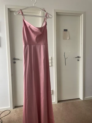 Rosa långklänning med smala axelband - Säljer en elegant rosa långklänning med smala axelband som knyts upptill. Klänningen har en enkel och stilren design med lätt utställd kjol och mjukt fall. Perfekt för dig som gillar en feminin och klassisk look. Den har en ragkedja på sidan och en slit i kjolen. Axel banden går att knyta själva och väls hur långa man vill ha dem. Den är knappt använd och har endast provats hemma då den var fel storlek. 